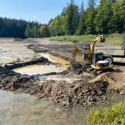 Żółta koparka i wywrotka pracują przy pogłębianiu i modelowaniu dna wyschniętego zbiornika wodnego, widoczne ślady świeżego wykopu i nagromadzona ziemia, w tle las.