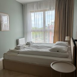 RUT-DOM Usługi Remontowo Wykończeniowe - Jasna sypialnia po remoncie: podwójne łóżko z pościelą, stolik nocny, okno z firanami i zasłonami. Ściana w odcieniach zieleni, obraz na ścianie, drewniana podłoga.