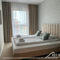 RUT-DOM Usługi Remontowo Wykończeniowe - Jasna sypialnia z dwoma łóżkami, beżowym zagłówkiem i wzorzystą tapetą. Widok na okno z zasłonami. Minimalistyczny design z akcentami kolorystycznymi w postaci poduszek.