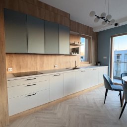 RUT-DOM Usługi Remontowo Wykończeniowe - Nowoczesna kuchnia z drewnianą ścianą, szarymi szafkami i białymi frontami. Podłoga w jodełkę, okno z widokiem na budynek. Minimalistyczna lampa sufitowa.