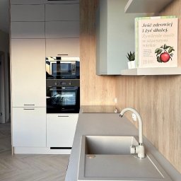 RUT-DOM Usługi Remontowo Wykończeniowe - Jasna kuchnia po remoncie: zabudowa z piekarnikiem i mikrofalówką, szary blat z zlewem, drewnopodobne panele ścienne, półka z książką o zdrowym odżywianiu.