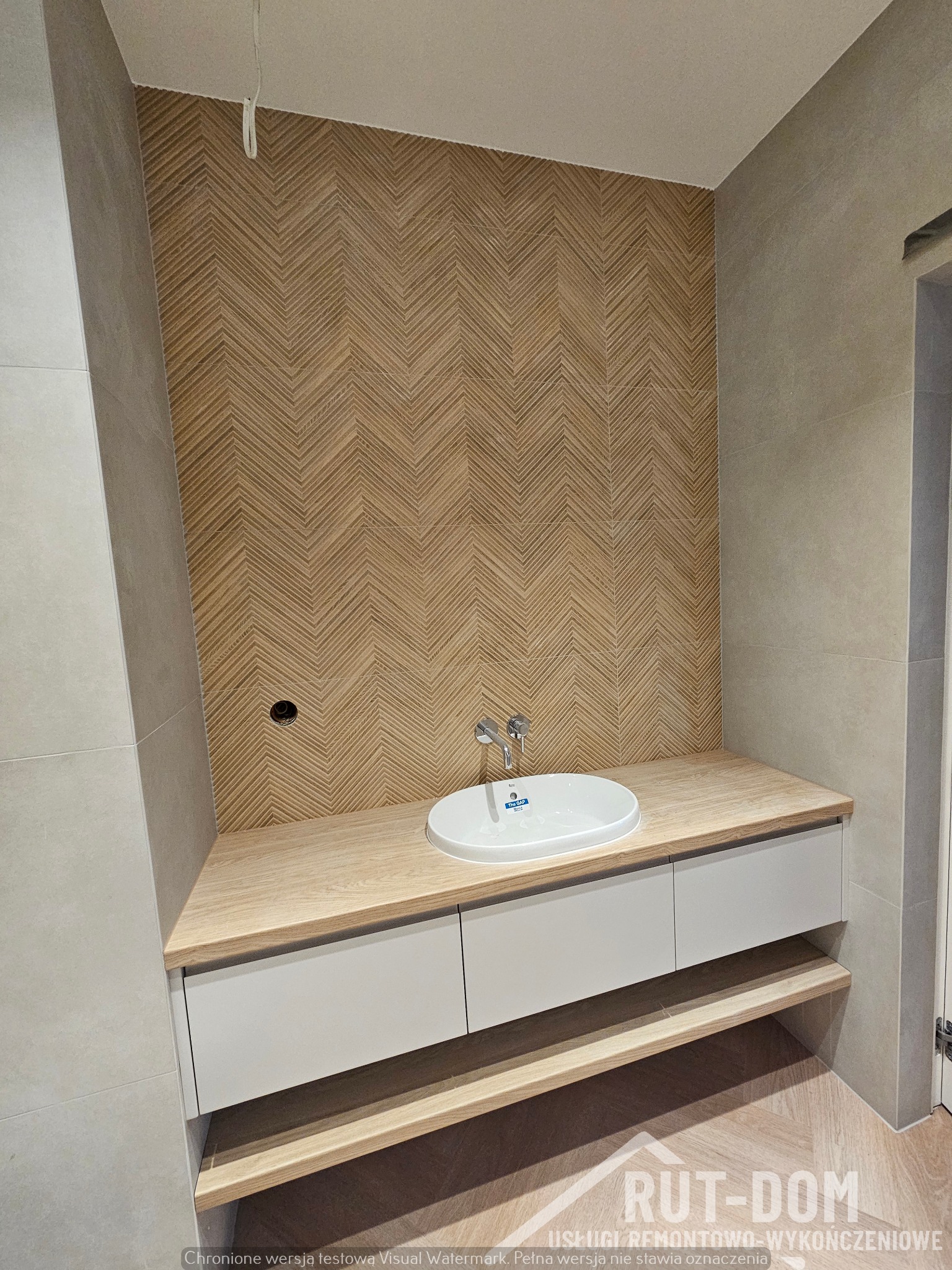 Nowoczesna łazienka z umywalką nablatową, drewnianym blatem i ścianą wykończoną płytkami w jodełkę, szare płytki na pozostałych ścianach, widoczne niedociągnięcia przy suficie.