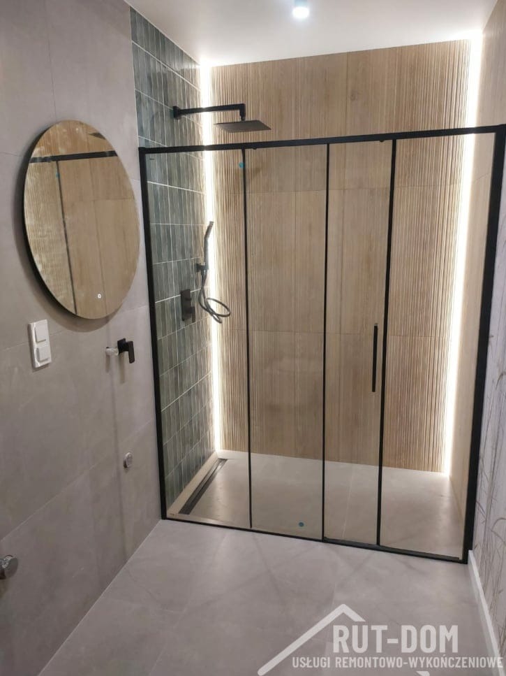 Nowoczesna kabina prysznicowa z czarnymi ramami i deszczownicą, oświetlona listwami LED, obok okrągłe lustro z podświetleniem na szarej ścianie z płytek. Uwagę zwraca dekoracyjna ściana z beżowych...