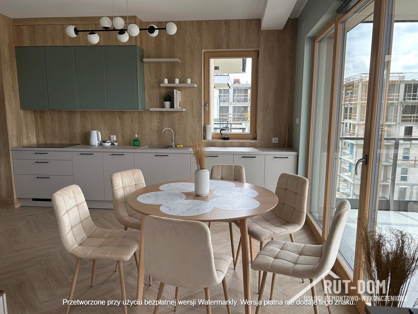 Jasna kuchnia z drewnianymi akcentami: okrągły stół z krzesłami, szare szafki i drewniana ściana. Widok z okna na budowę. Nowoczesne wykończenie.