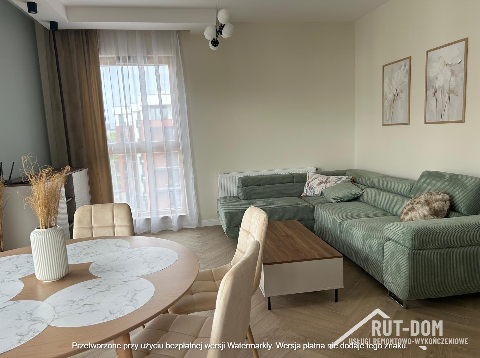 Jasne, przestronne wnętrze po remoncie: zielony narożnik, okrągły stół z dekoracją, parkiet w jodełkę. Dwa obrazy na ścianie. Widok z okna na budynek.
