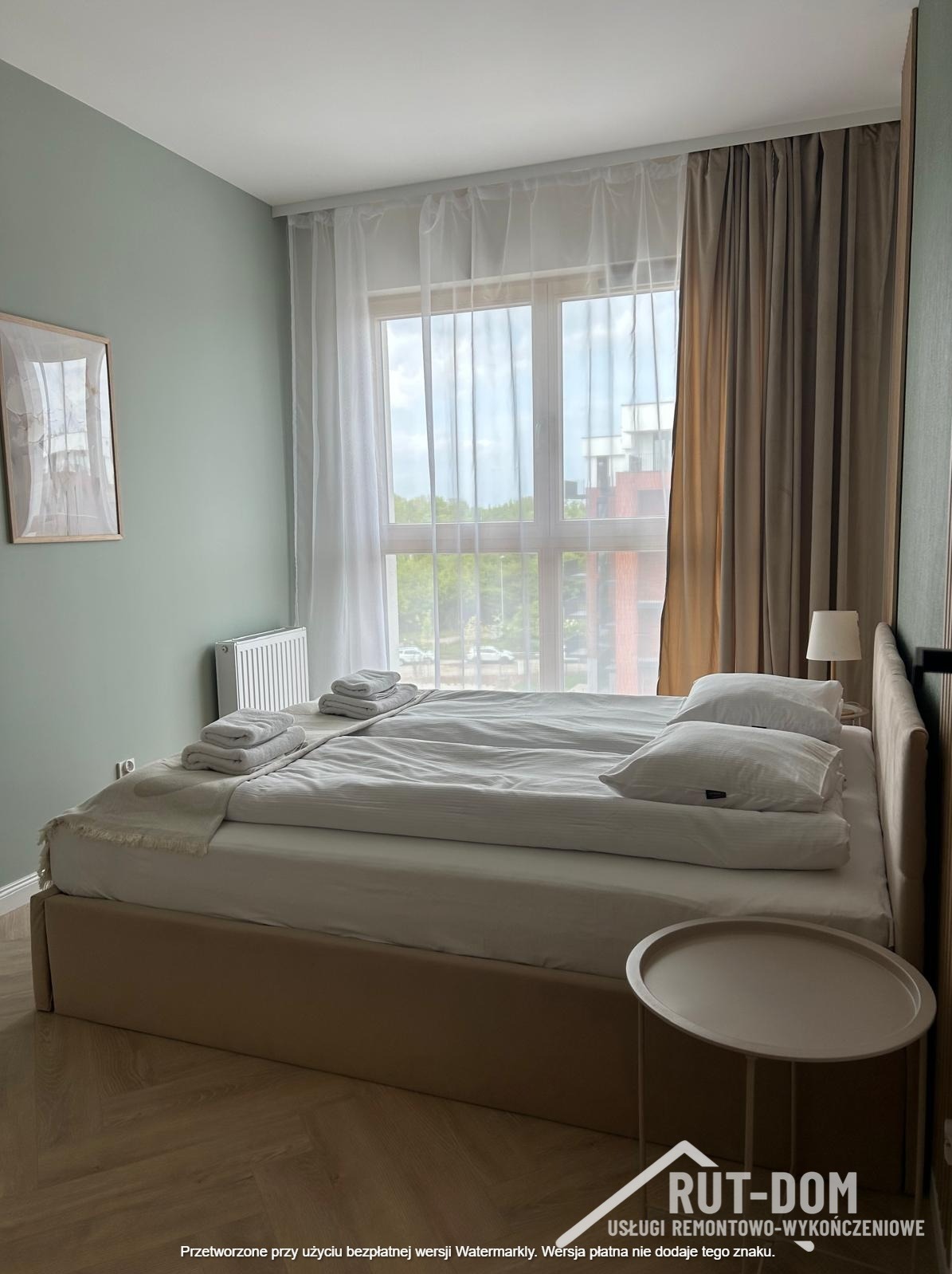 Jasna sypialnia po remoncie: podwójne łóżko z pościelą, stolik nocny, okno z firanami i zasłonami. Ściana w odcieniach zieleni, obraz na ścianie, drewniana podłoga.