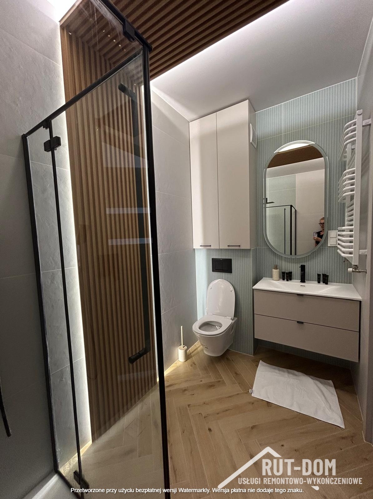 Nowoczesna łazienka z kabiną prysznicową typu walk-in, drewnianym akcentem na suficie i ścianie, oraz szafką z umywalką. Parkiet ułożony w jodełkę.