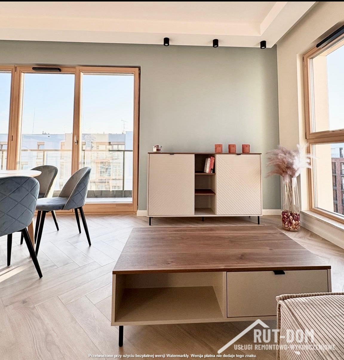 Jasne wnętrze z parkietem w jodełkę: stół, krzesła, komoda z dekoracjami i duże okna balkonowe. Ściana w kolorze pistacjowym. Nowoczesny design.