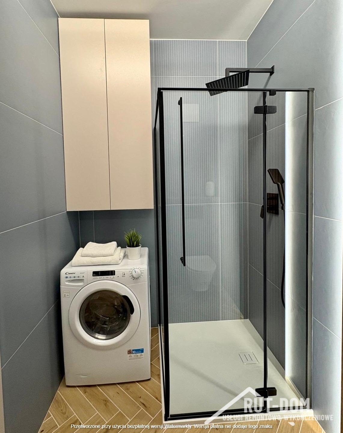 Nowoczesna łazienka z kabiną prysznicową typu walk-in z czarnymi profilami, pralką Candy i szafką wiszącą w beżowym odcieniu. Szare płytki na ścianach i drewnopodobna podłoga.