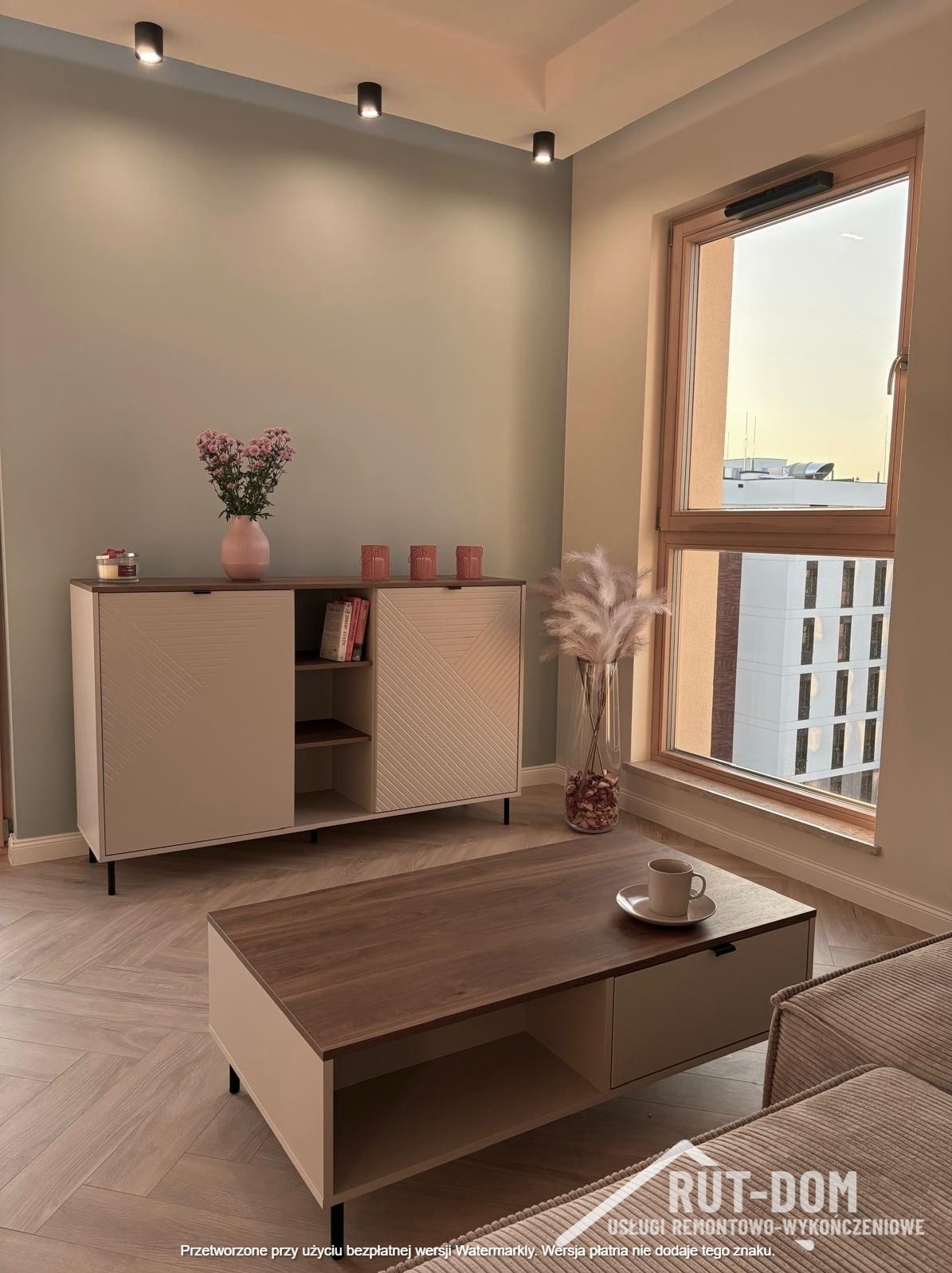 Jasne wnętrze po remoncie: komoda z dekoracjami, stolik kawowy z filiżanką, okno z widokiem na miasto i parkiet w jodełkę. Minimalistyczny design i stonowana kolorystyka.