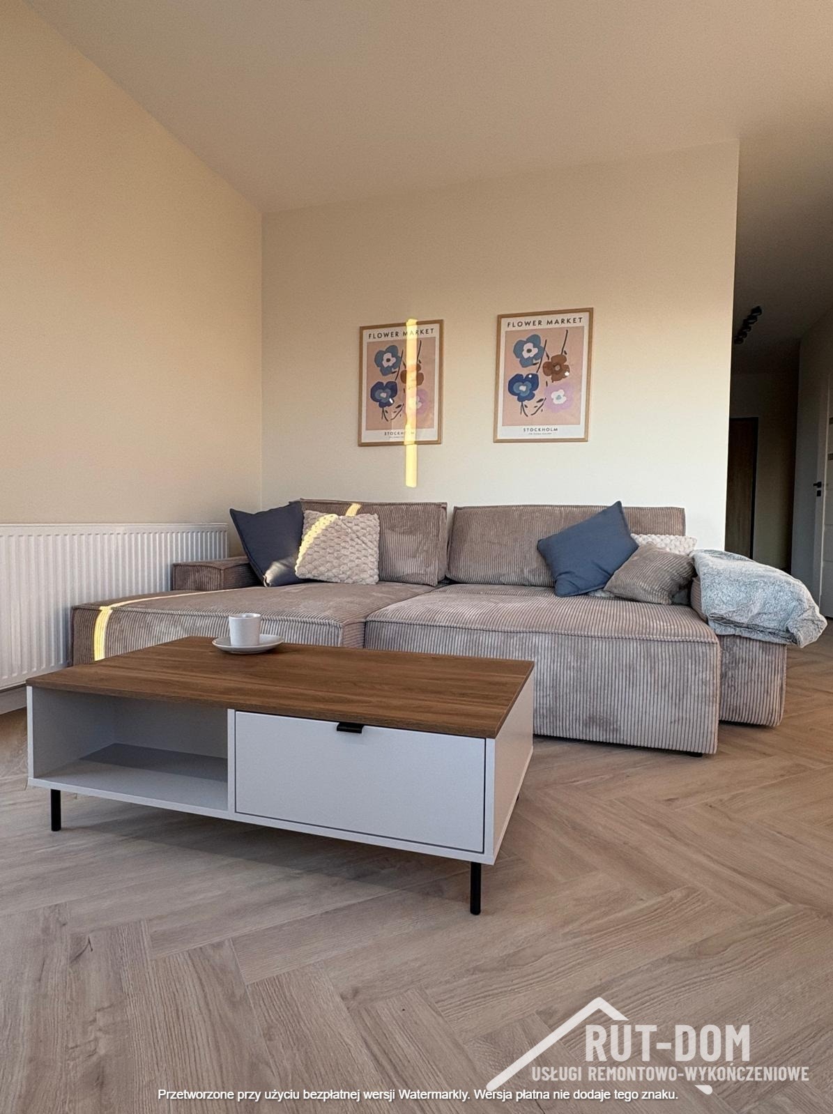 Jasny salon z parkietem w jodełkę, beżową sofą narożną, stolikiem kawowym z szufladą i dekoracjami ściennymi w postaci plakatów w ramkach.