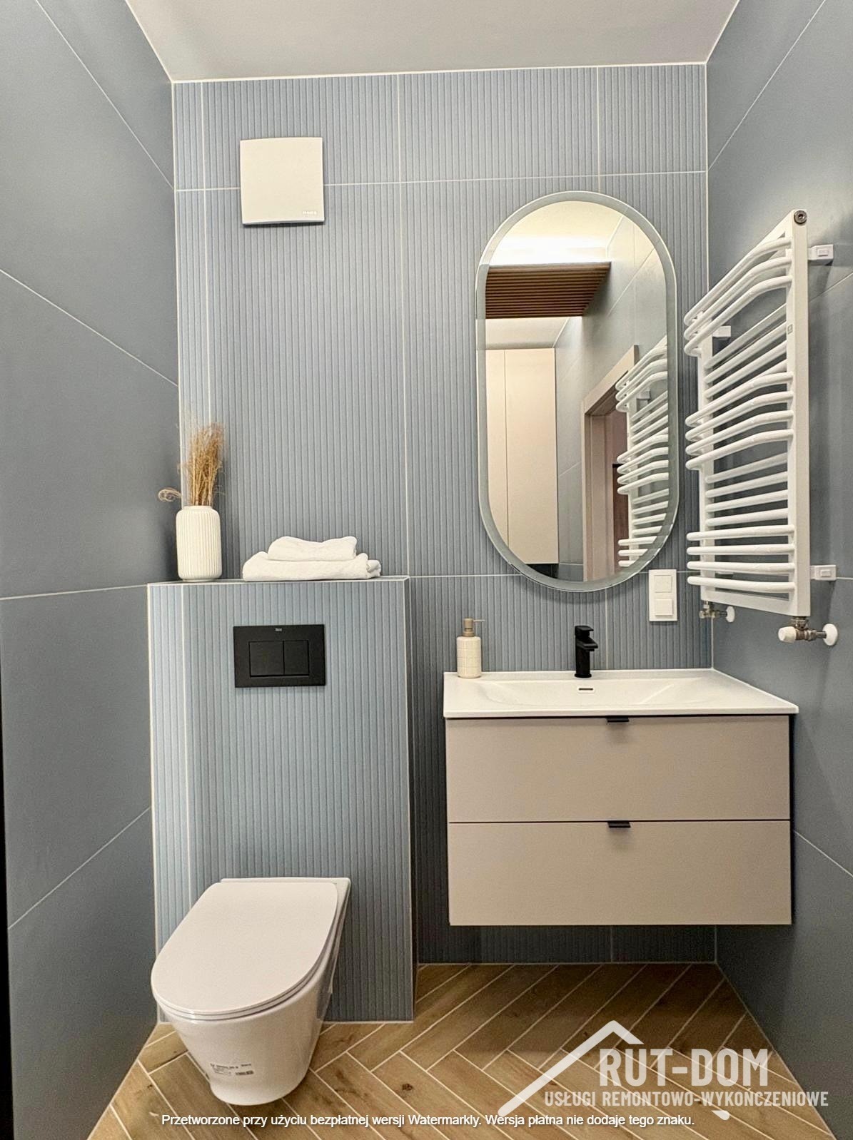 Niebieska łazienka z płytkami w paski, owalnym lustrem i drewnianą podłogą w jodełkę. W tle białe drzwi i grzejnik drabinkowy.