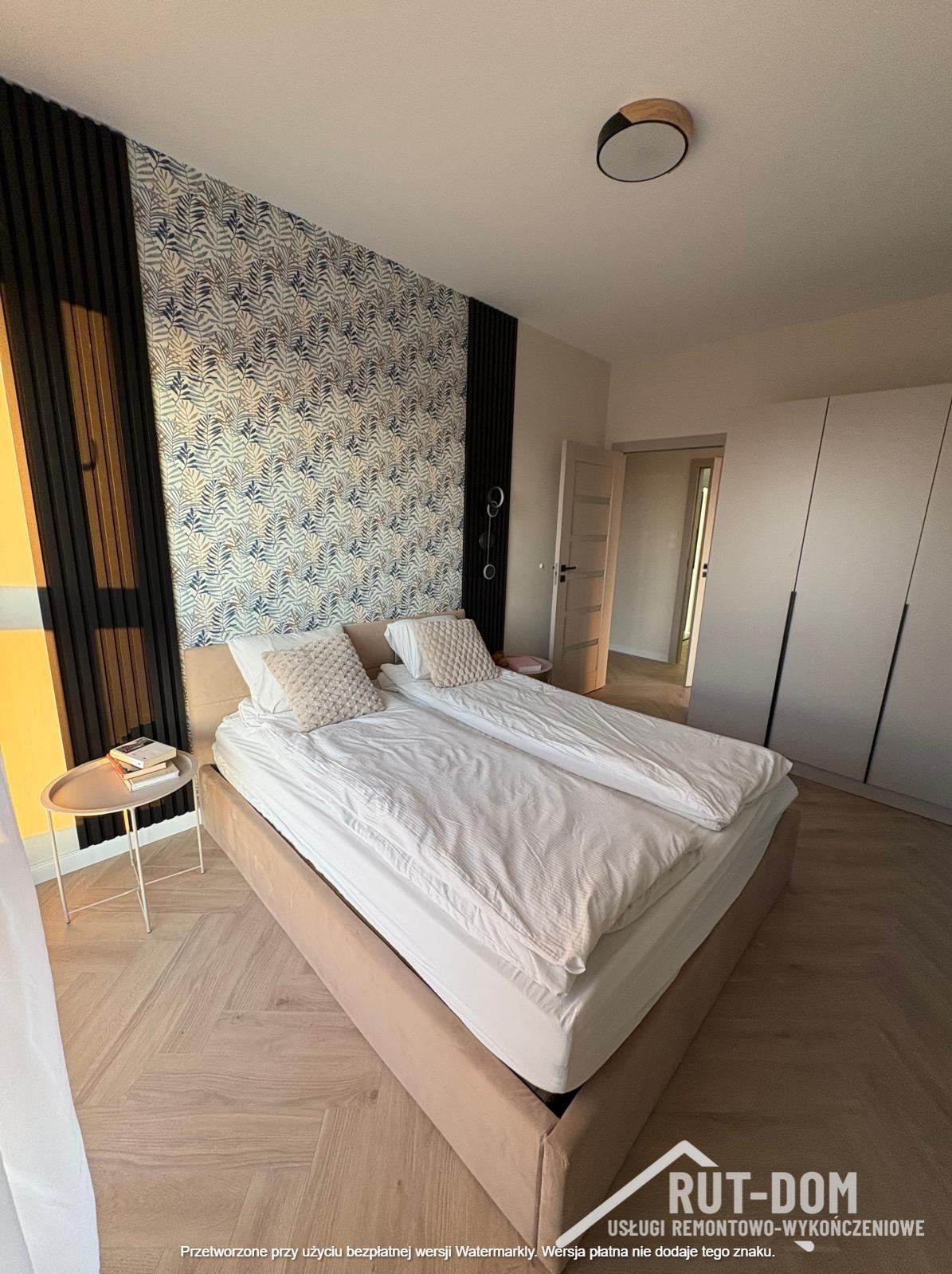 Nowoczesna sypialnia z łóżkiem małżeńskim, wzorzystą tapetą w liście i drewnianymi lamelami. Parkiet w jodełkę, szafa w zabudowie i okrągła lampa sufitowa.