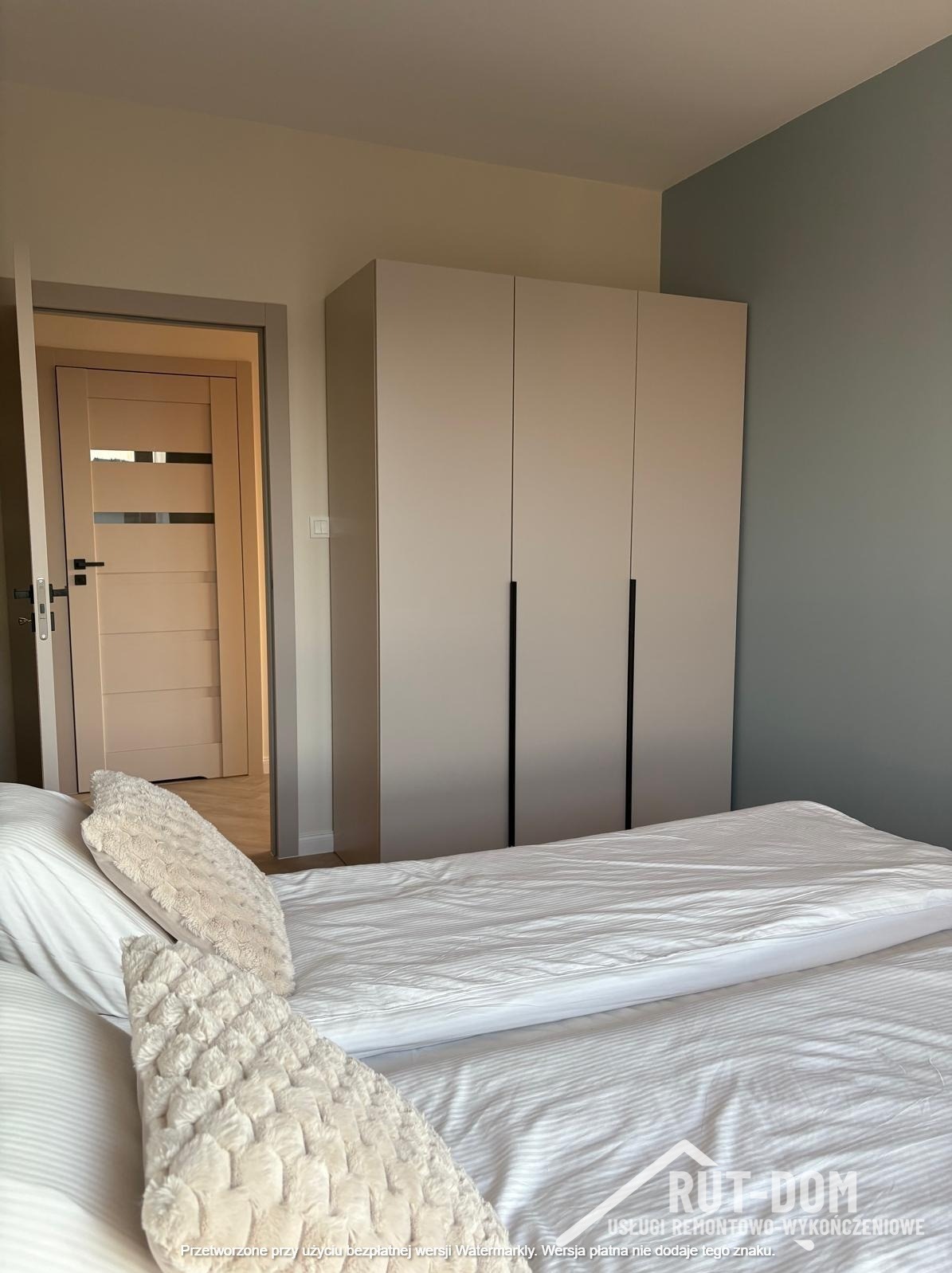 Beżowa szafa wnękowa z czarnymi uchwytami w sypialni, widok z perspektywy łóżka. Ściana w odcieniach beżu i zieleni. Widoczny fragment otwartych drzwi.