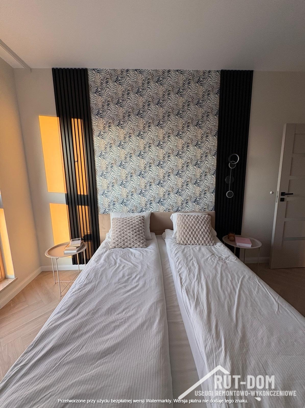 Sypialnia z łóżkiem, tapetą w liście, panelami ściennymi i stolikami nocnymi. Widoczne elementy wykończenia wnętrz w nowoczesnym stylu.