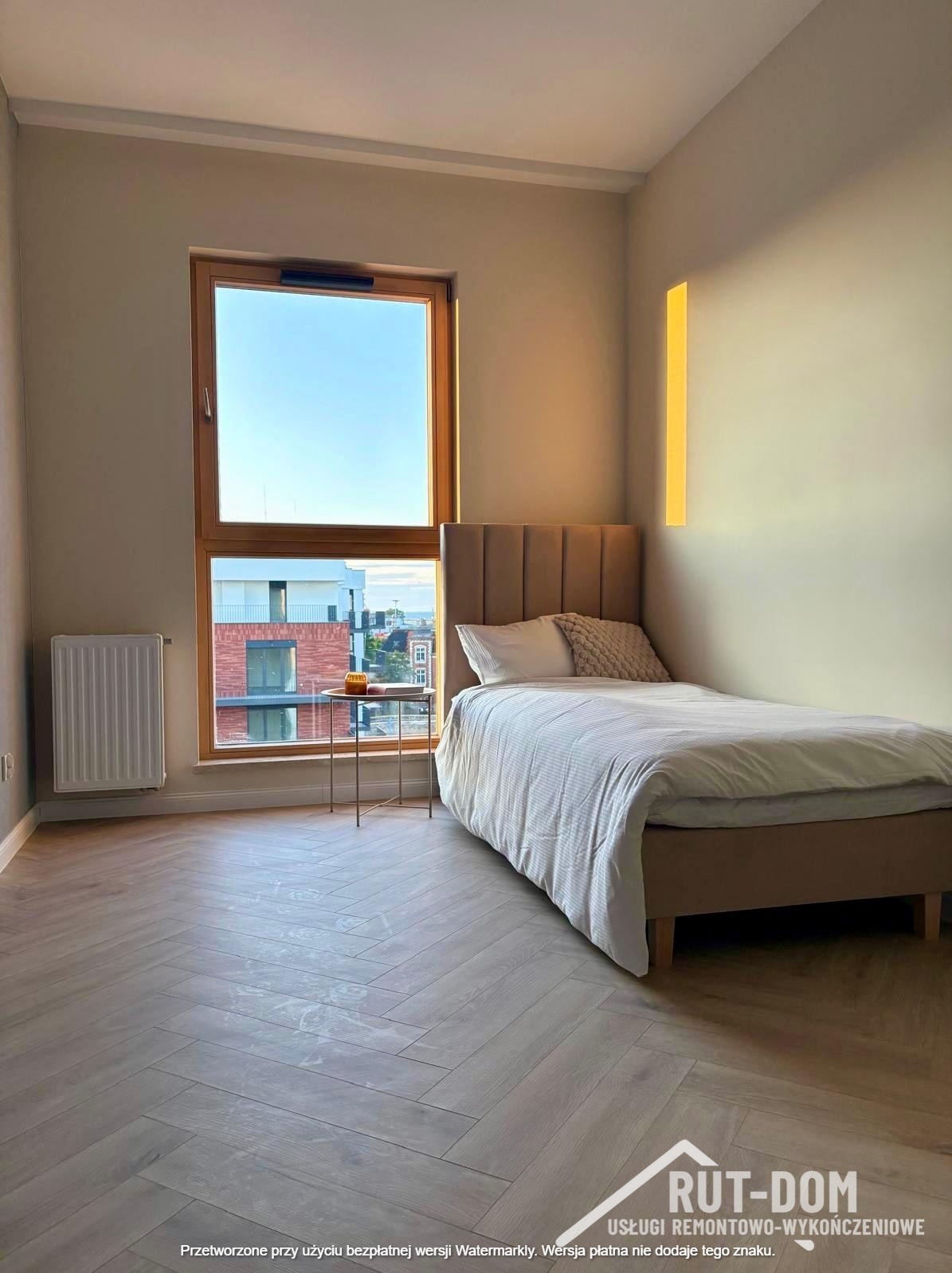 Jasny pokój po remoncie: parkiet w jodełkę, beżowe ściany, okno z widokiem na miasto. Minimalistyczny wystrój, łóżko z beżowym wezgłowiem i mały stolik.