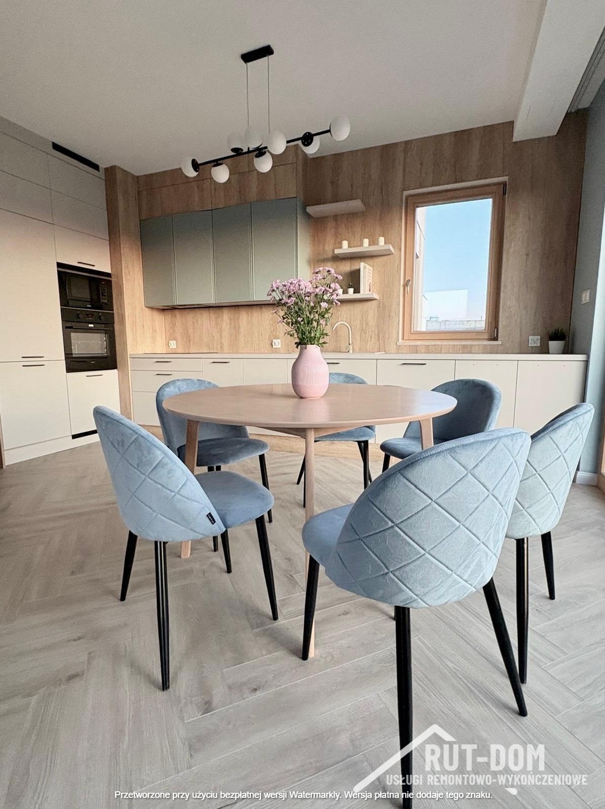 Jasna kuchnia z okrągłym stołem i błękitnymi krzesłami z pikowaniem. Na stole wazon z kwiatami. Parkiet ułożony w jodełkę. Widoczne zabudowy kuchenne i okno.