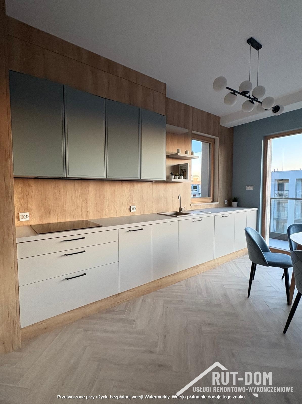 Nowoczesna kuchnia z drewnianą ścianą, szarymi szafkami i białymi frontami. Podłoga w jodełkę, okno z widokiem na budynek. Minimalistyczna lampa sufitowa.