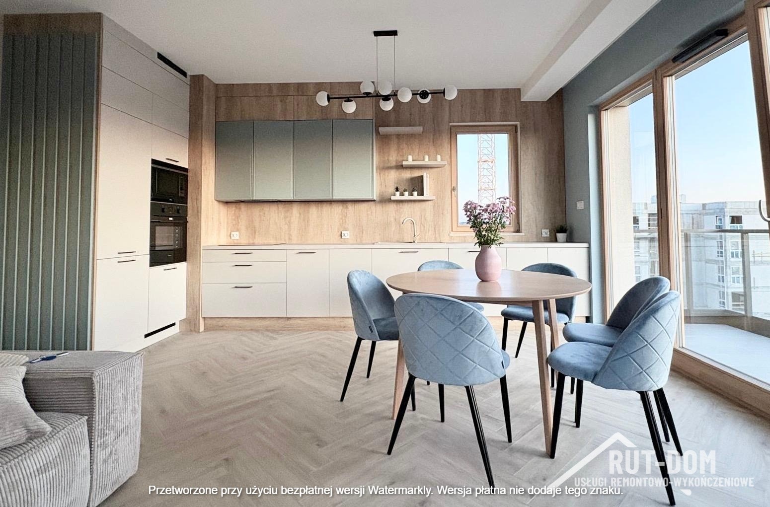 Jasna kuchnia z drewnianymi akcentami, okrągłym stołem i błękitnymi krzesłami. Widok na miasto przez duże okno. Nowoczesne wykończenie wnętrz.