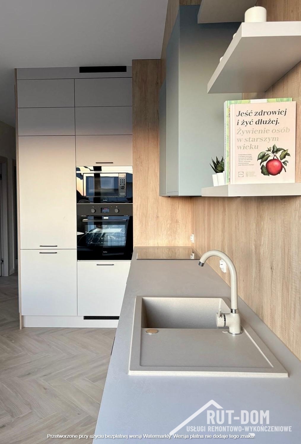 Jasna kuchnia po remoncie: zabudowa z piekarnikiem i mikrofalówką, szary blat z zlewem, drewnopodobne panele ścienne, półka z książką o zdrowym odżywianiu.