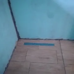 Niebieski odpływ liniowy wkomponowany w beżowe płytki podłogowe, ściany pomalowane na turkusowo, widoczne niedociągnięcia przy łączeniu ścian i podłogi.