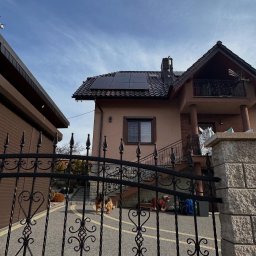 Green-Tech Sp. z o.o. - Dom jednorodzinny z zainstalowanymi panelami słonecznymi na dachu, widok zza ozdobnego, kutego ogrodzenia; na podjeździe pies rasy owczarek niemiecki.