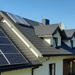 Panele słoneczne na dachu domu jednorodzinnego z czarną dachówką, widoczne góry w tle i logo firmy Green Tech w prawym górnym rogu.