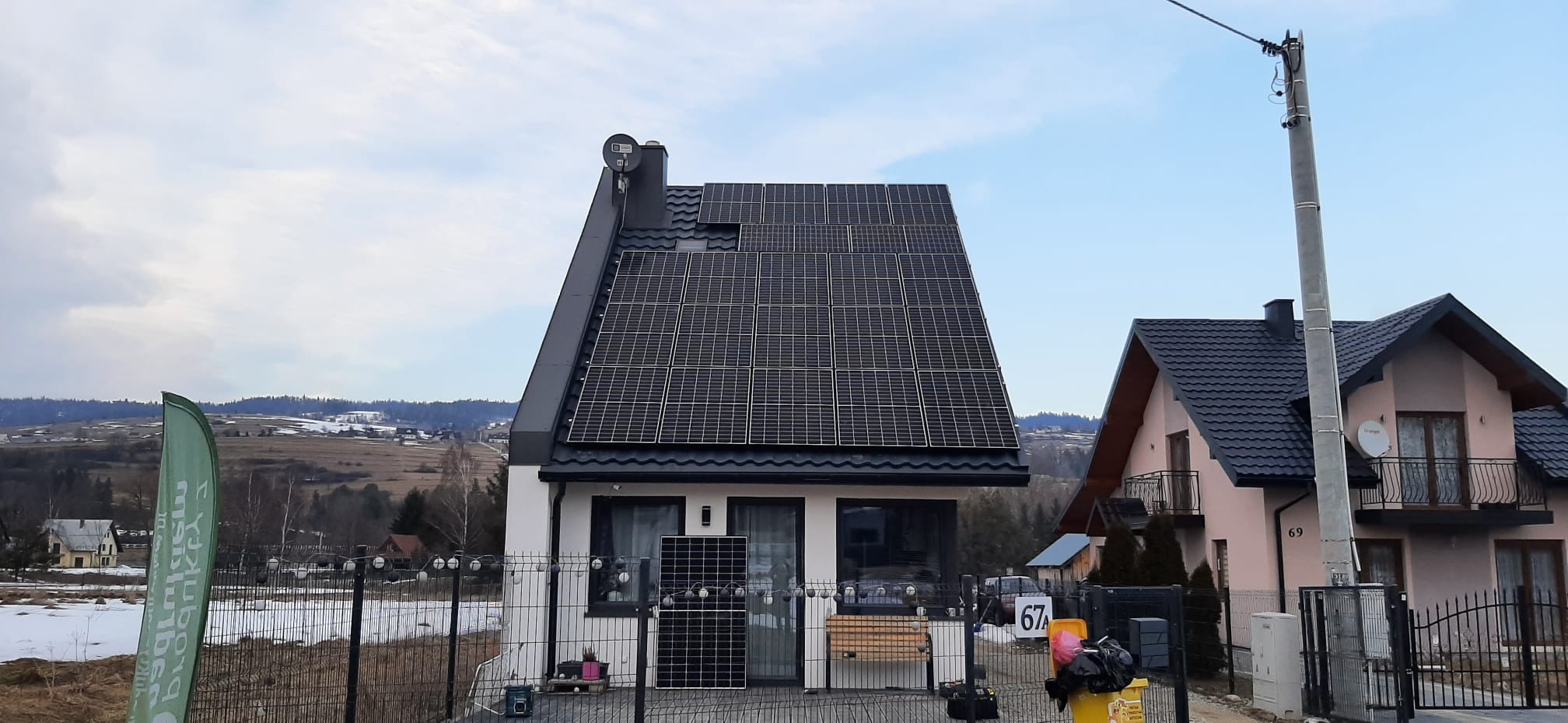 Dom jednorodzinny z panelami słonecznymi na dachu, widoczne panele przygotowane do montażu na ziemi, w tle pagórkowaty krajobraz i częściowo ośnieżone pola.