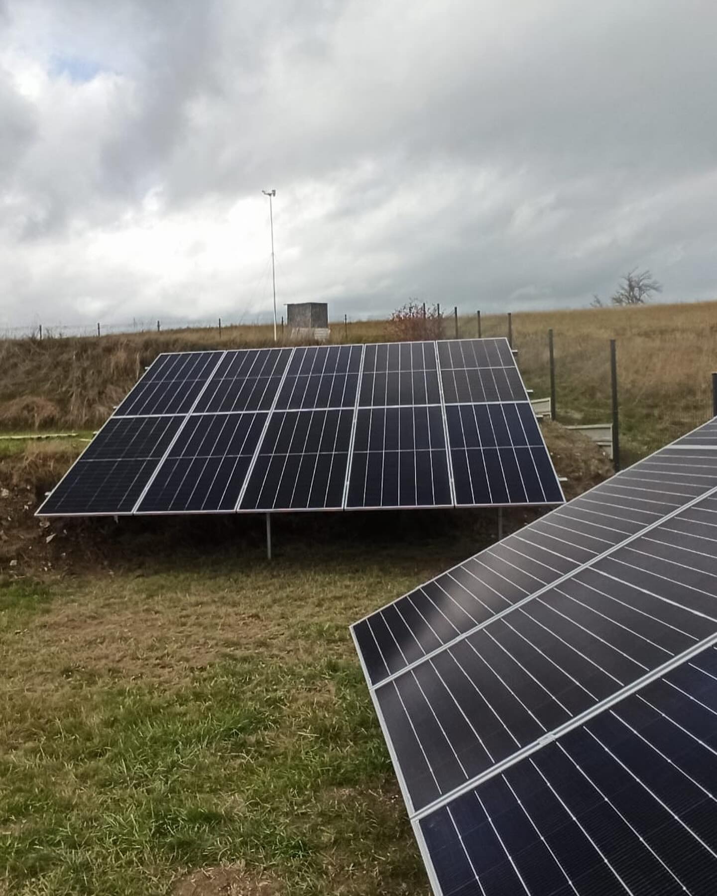 Dwa panele słoneczne ustawione na trawiastym wzgórzu pod pochmurnym niebem, z widocznym w tle budynkiem i masztem meteorologicznym.