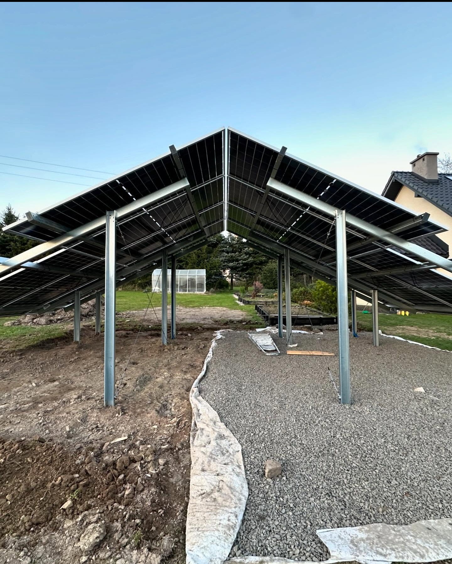 Konstrukcja wsporcza pod panele słoneczne w trakcie montażu, widok od dołu z perspektywą na ogród i szklarnię w tle.