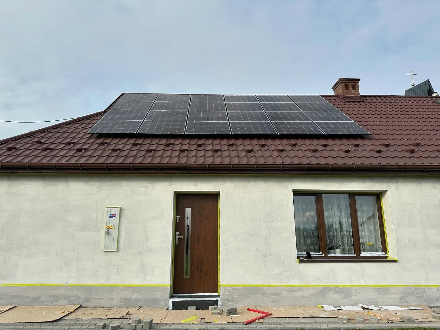 Panele słoneczne zamontowane na brązowym dachu domu jednorodzinnego z beżową elewacją, widoczne również okno z zasłonami i brązowe drzwi wejściowe, na ziemi kostka brukowa i narzędzia.