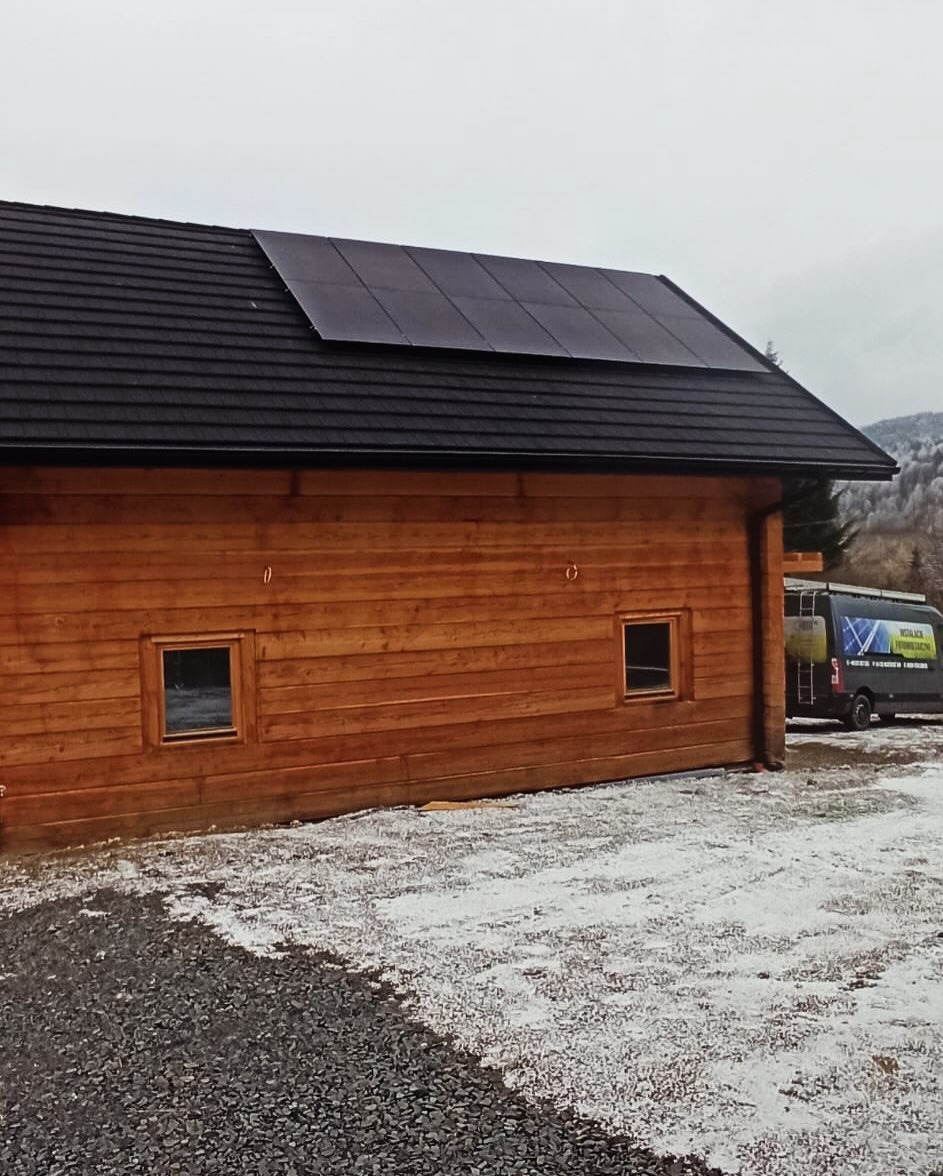 Drewniany budynek z panelami słonecznymi na dachu, widoczny samochód serwisowy w tle, teren pokryty śniegiem.
