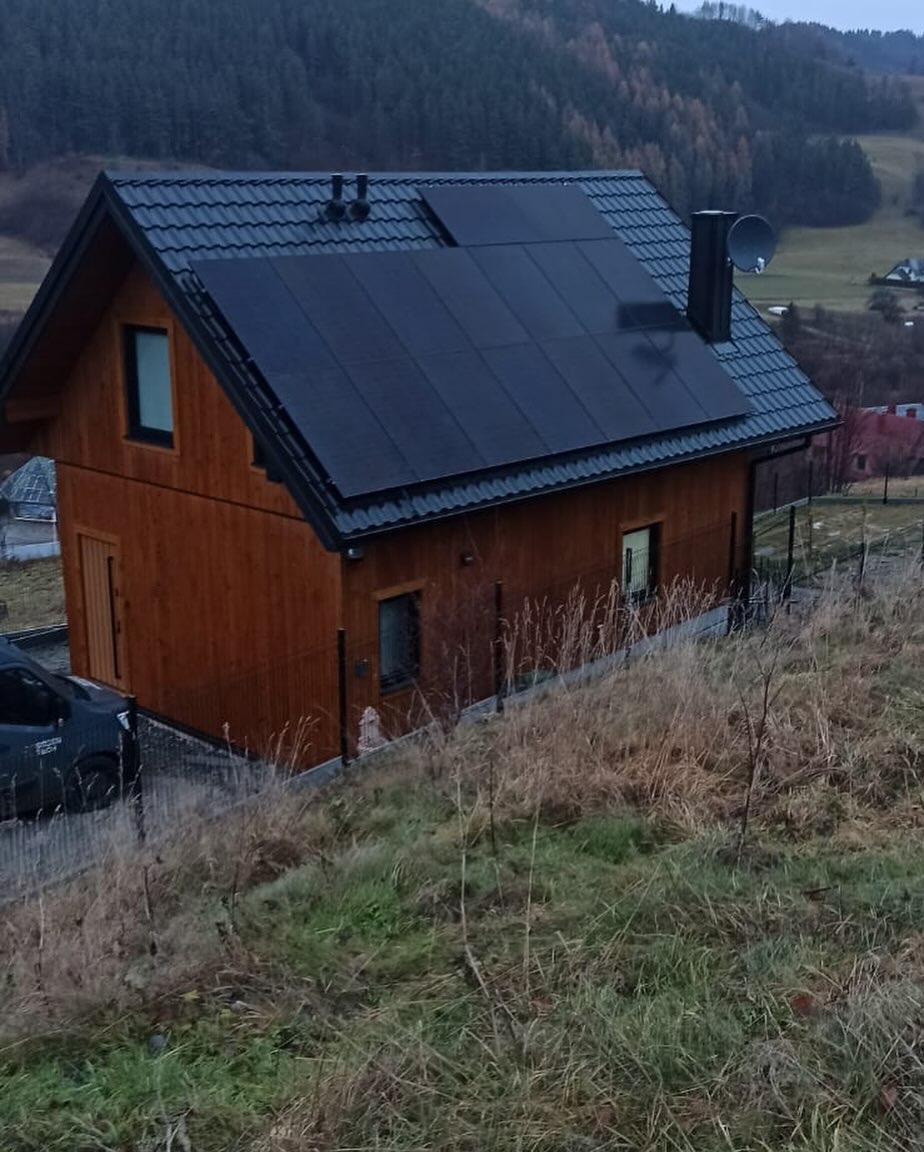 Drewniany dom z czarnymi panelami słonecznymi na dachu, kominem i anteną satelitarną, widok z lekko podwyższonej perspektywy na tle zalesionych wzgórz.