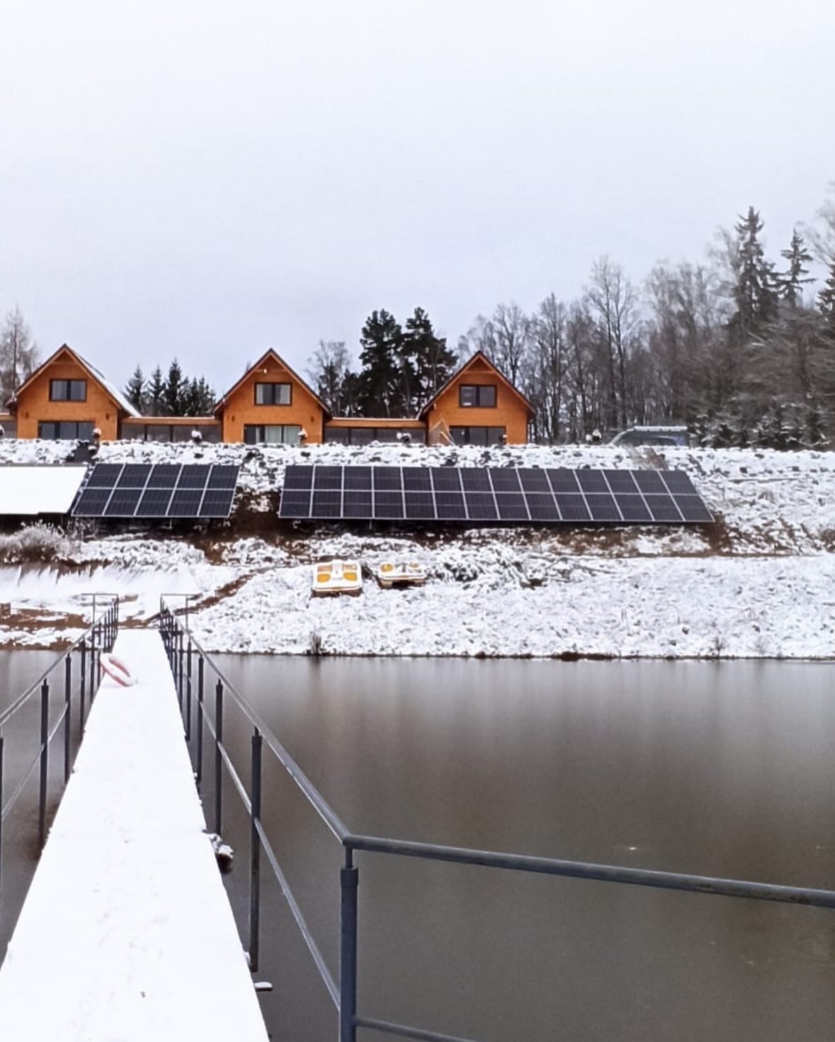 Zimowy krajobraz z trzema domami z panelami słonecznymi na dachu, widok z pomostu nad wodą, na śnieżnym wzgórzu.