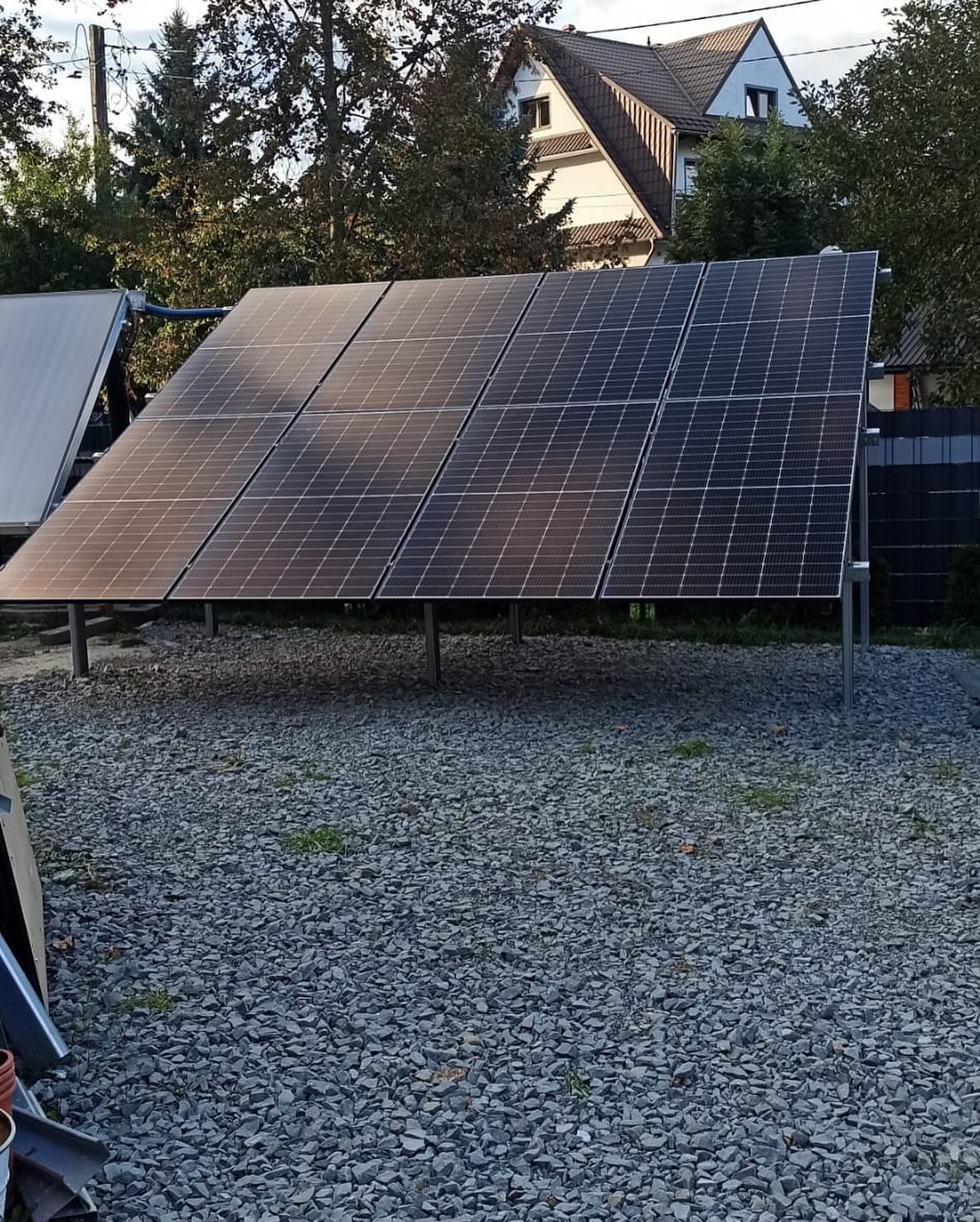Panel fotowoltaiczny na metalowej konstrukcji, ustawiony na żwirowym podłożu, w tle dom jednorodzinny z brązowym dachem.