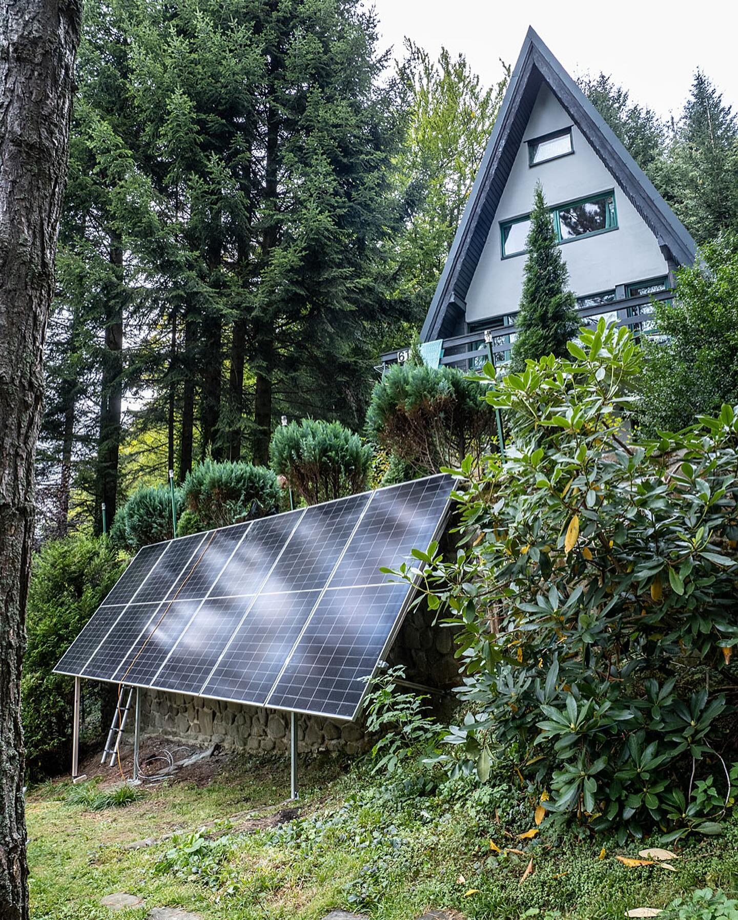 Panel słoneczny zamontowany na kamiennej podstawie przed domem w stylu A-frame otoczonym drzewami iglastymi i zielenią.