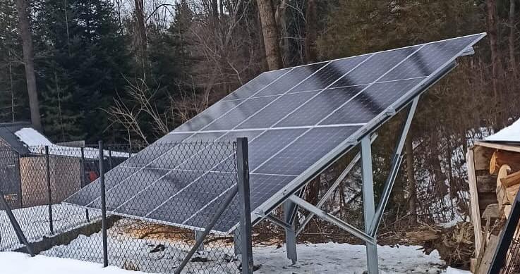 Instalacja fotowoltaiczna na metalowej konstrukcji w zimowym krajobrazie, widoczny fragment ogrodzenia z siatki i składowane drewno.