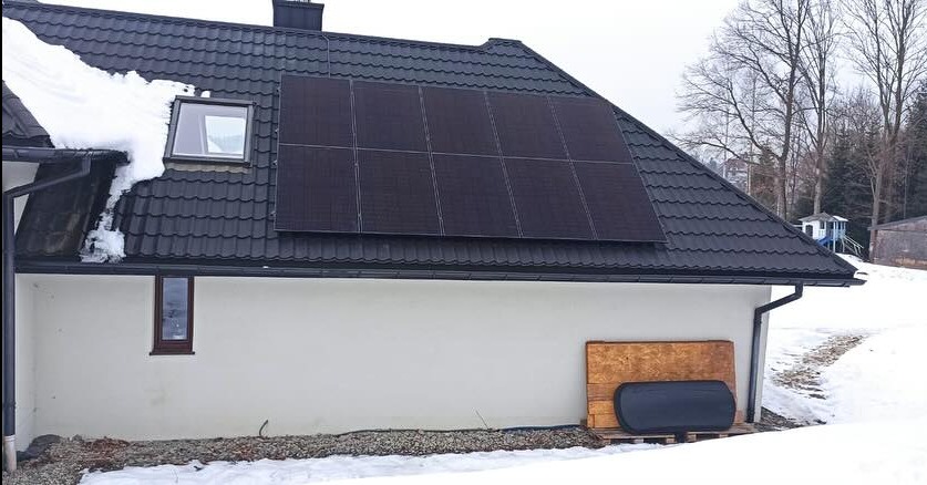Czarny panel fotowoltaiczny na dachu domu pokrytym ciemną dachówką, z resztkami śniegu na dachu i w otoczeniu.
