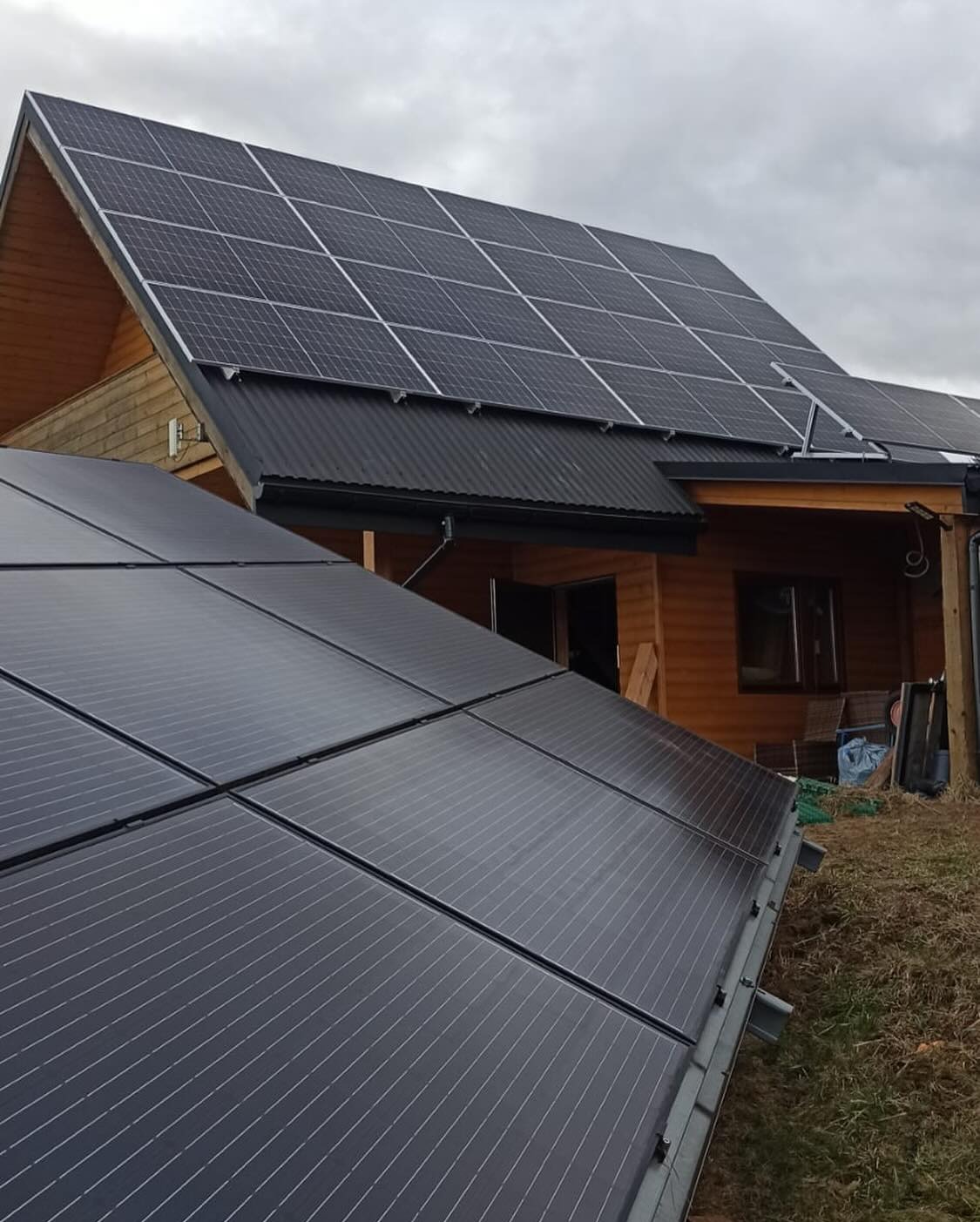 Panele słoneczne na dachu drewnianego domu jednorodzinnego, widoczne mocowania i fragment instalacji na ziemi.