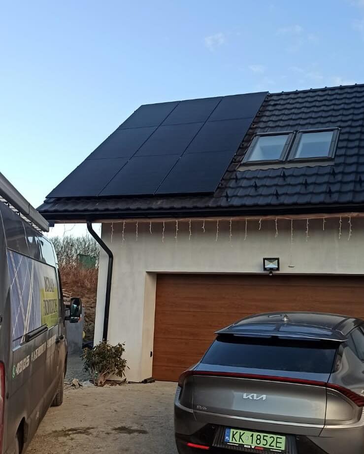 Panele słoneczne na dachu domu jednorodzinnego z oknami dachowymi, zaparkowany samochód elektryczny Kia EV6 i bus firmy instalacyjnej z widocznym numerem telefonu.
