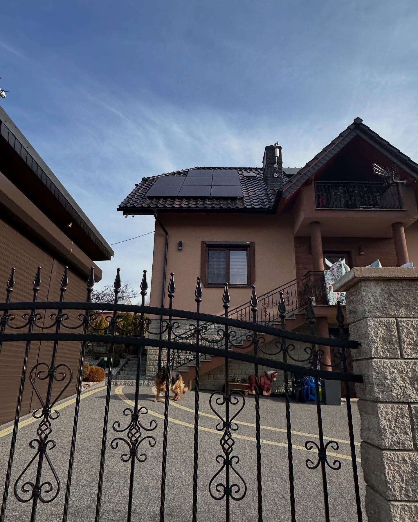 Dom jednorodzinny z zainstalowanymi panelami słonecznymi na dachu, widok zza ozdobnego, kutego ogrodzenia; na podjeździe pies rasy owczarek niemiecki.