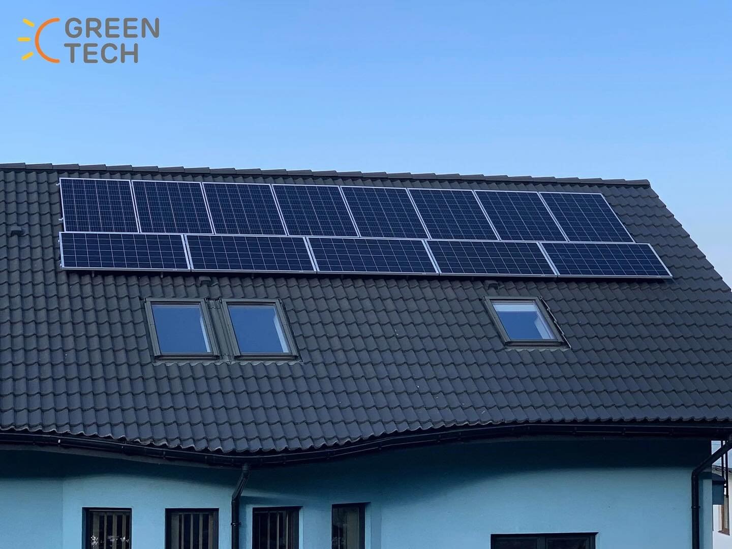 Panele słoneczne na ciemnym dachu domu z trzema oknami dachowymi, widoczne logo 'GREEN TECH' w górnym lewym rogu.