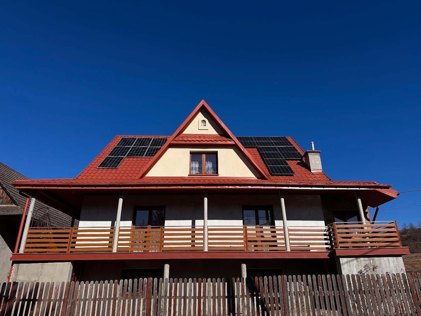 Dwukondygnacyjny dom z czerwonym dachem i panelami słonecznymi pod błękitnym niebem, widziany zza drewnianego płotu i balkonu.