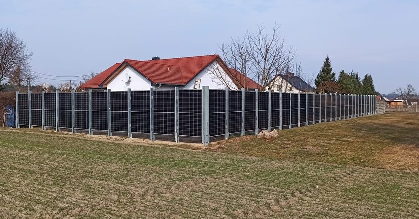Długa instalacja fotowoltaiczna pełniąca funkcję ogrodzenia, zamontowana na metalowych słupkach, z domami w tle i nieprzygotowanym terenem przed nią.