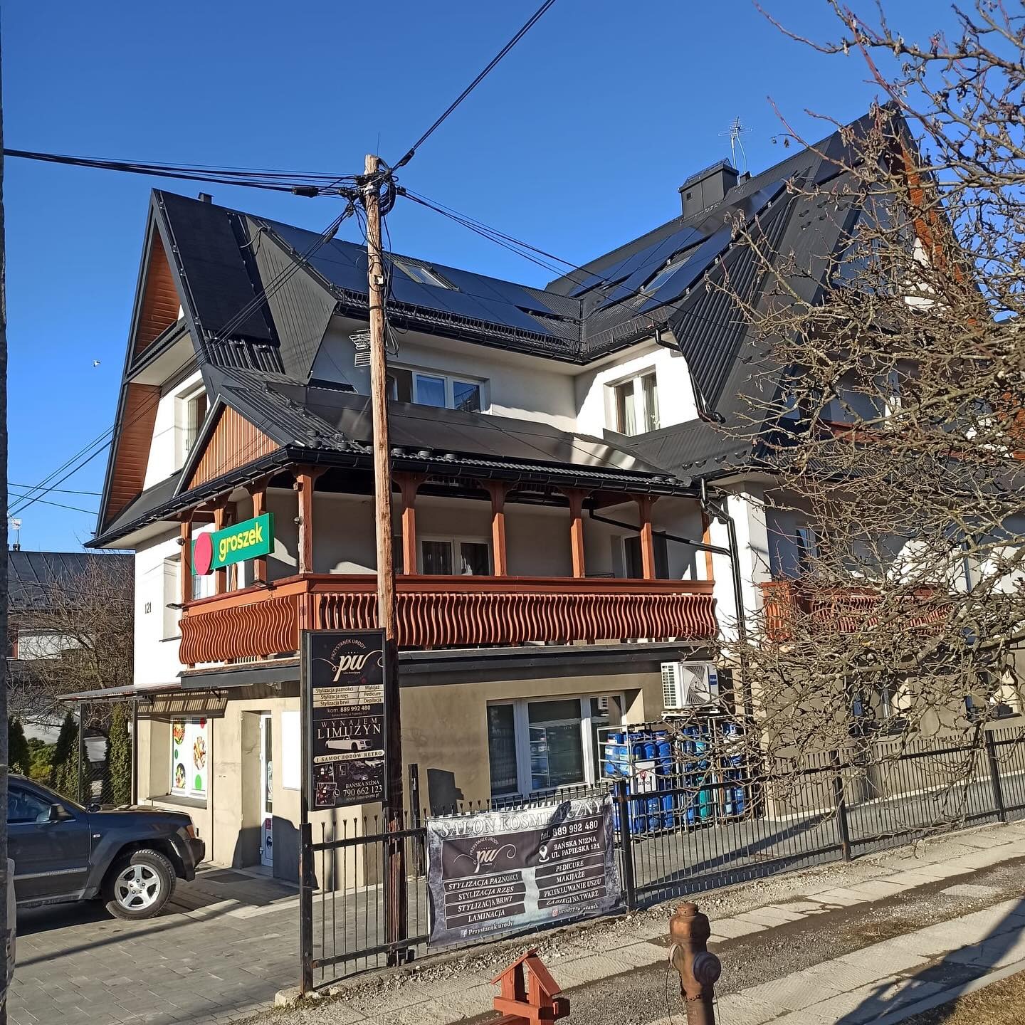 Dwupiętrowy budynek z czarnym dachem, na którym zamontowane są panele słoneczne. Na parterze sklep 'Groszek' i salon kosmetyczny. Słoneczny dzień.