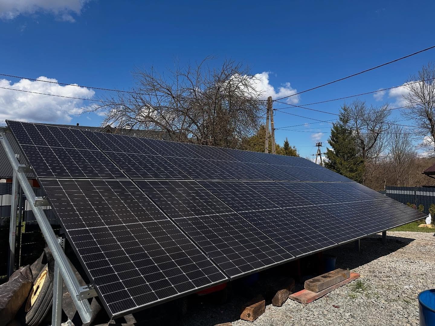 Instalacja paneli słonecznych na metalowej konstrukcji, widoczne elementy montażowe i otoczenie posesji z drzewami i niebem.