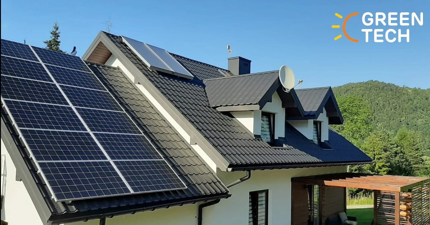 Panele słoneczne na dachu domu jednorodzinnego z czarną dachówką, widoczne góry w tle i logo firmy Green Tech w prawym górnym rogu.
