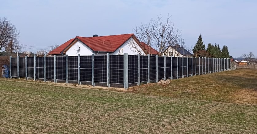 Długa instalacja paneli słonecznych, pełniąca funkcję ogrodzenia, zamontowana na metalowych słupkach, na tle domów jednorodzinnych i pola.