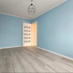 Dam-bud - Jasne, błękitne ściany w pokoju z szarymi panelami podłogowymi i nowoczesną lampą wiszącą, białe drzwi z przeszkleniami po lewej stronie, widok na kolejne pomieszczenie.