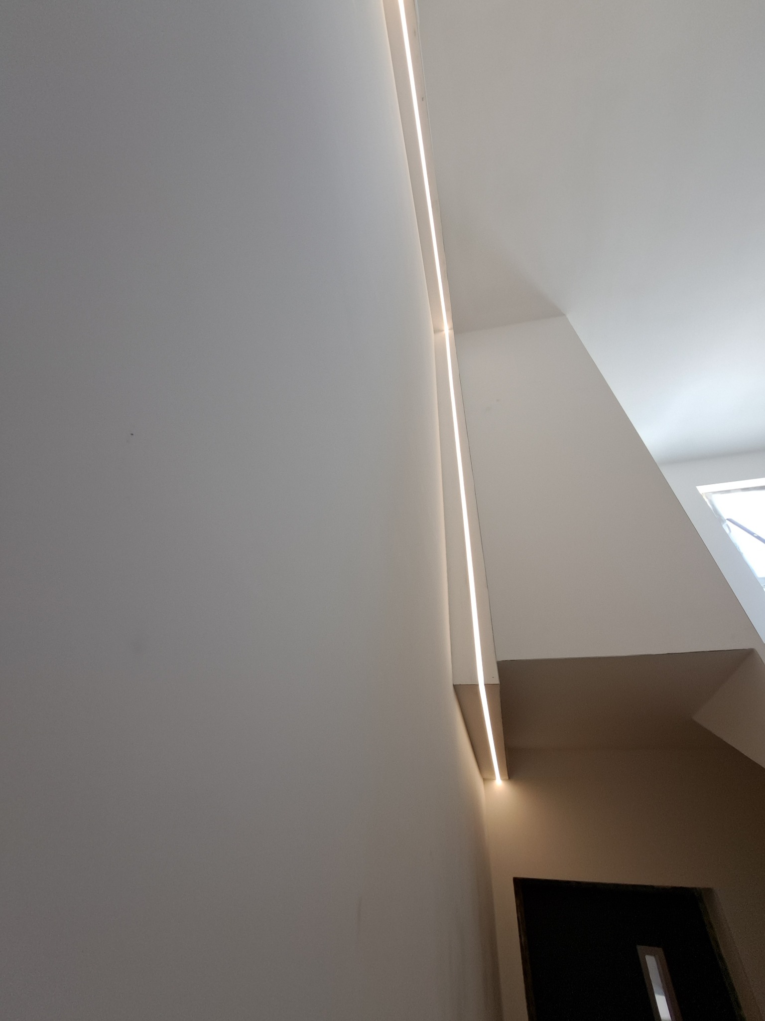 Nowoczesne oświetlenie LED wpuszczone w ścianę, tworzące pionową linię światła w korytarzu z białymi ścianami i skośnym sufitem.
